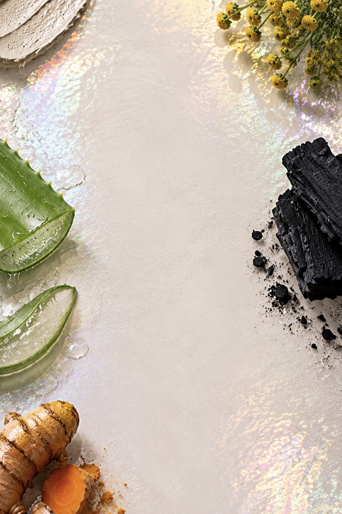 Strange ingredients including charcoal, turmeric, blue tansy flowers and aloe vera arranged around a soft pearlescent surface